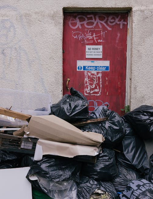 A collection of black plastic rubbish bags and discarded cardboard and packaging materials are piled in front of a red metal door set into a beige, textured wall. The door features graffiti markings in white and pink spray paint, including some tags and illegible characters, with multiple stickers attached. One prominent white sticker reads 'NO MONEY' and 'KEEP CLEAR' in blue, and another smaller sticker displays 'NO PARKING' text. The rubbish bags appear loosely tied, with some leaning against the door, and the various materials include crumpled cardboard sheets and plastic wrapping. The area looks like an outdoor urban setting, possibly a back alley or service access point, where private waste collection or on-site clearance might be taking place, illustrating a typical scene for independent rubbish disposal services like those provided by Rubbish Removal Ealing. The natural lighting accentuates the textures of the trash and the weathered surface of the door, emphasizing the contrast between the rubbish and the surrounding environment.