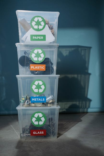 A set of four transparent plastic recycling bins stacked vertically against a wall, each labeled with a different material category: the top bin for paper, the second for plastic, the third for metals, and the bottom for glass. The paper bin contains a few crumpled sheets of paper, the plastic bin holds various plastic bottles and containers, the metals bin includes aluminium cans and metal scraps, and the glass bin contains glass bottles and jars. Each bin features a green recycling symbol on the front, with colour-coded labels in white, orange, blue, and red respectively, clearly indicating the material type for waste separation. The bins are placed in a light, indoor environment, with natural or artificial lighting illuminating the scene and casting subtle shadows on the floor. This setup reflects typical waste sorting and recycling practices that support efficient rubbish management and align with alternative rubbish removal methods, such as independent collection or on-site waste segregation, observed in household or community settings. The background includes a portion of a grey storage unit or cabinet, and the scene exemplifies organized waste disposal relevant to private or non-local-authority rubbish services, as highlighted by Rubbish Removal Ealing.