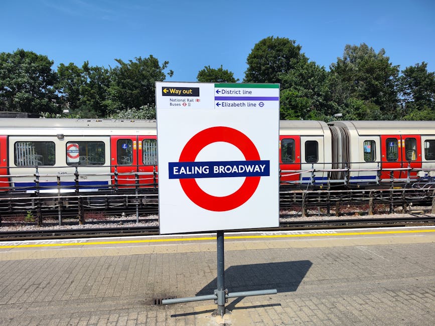 A photograph of a sign at Ealing Broadway railway station, featuring the red and white London Underground roundel with a blue band displaying the station name. The sign is mounted on a metal pole situated on a paved platform area. Behind the sign, there is a modern passenger train with a silver body accented by red and blue stripes running horizontally along its side. The train has multiple windows, some with visible reflections or partially open blinds, and is positioned on railway tracks that run parallel to the platform. In the background, there are lush green trees with dense foliage under a clear blue sky, indicating a sunny day. The surrounding environment suggests a typical urban railway station setting, with safety barriers and signals visible nearby, supporting efficient passenger movement and alternative transportation options outside of the main public transport system.