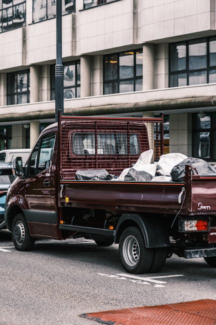 A small dark red flatbed truck parked on an urban street in front of a modern office building with large glass windows and a concrete facade. The truck's open cargo area is filled with various types of waste, including white and black plastic bags and loose debris, indicating recent rubbish collection or disposal. The vehicle is positioned close to the curb, with a traffic sign and part of a white van visible nearby. The scene is lit by natural daylight, creating clear shadows on the pavement. The overall environment suggests an area where independent rubbish removal or private waste handling services, such as those provided by Rubbish Removal Ealing, might operate, especially for local property clearances or commercial waste management.