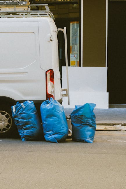 The image shows the rear side of a white delivery van parked on a street, with three large blue plastic rubbish bags placed on the pavement next to it. The bags appear full and are slightly crumpled, made from a glossy, durable material, with one bag leaning against the van's rear wheel and the other two positioned nearby. In the background, there is a modern building with a glass door and a white panel on the exterior wall, indicating a commercial or residential property. The scene is illuminated by natural daylight, with minimal shadows cast on the pavement, suggesting a clear or partly cloudy sky. This setting illustrates the context of private waste disposal, which can be an alternative to curbside collection, with Rubbish Removal Ealing potentially offering services that facilitate on-site rubbish clearance without reliance on local authority pickup. The focus on the bags' placement next to the vehicle highlights an aspect of independent waste handling relevant to fast, same-day rubbish removal services in the Ealing area.