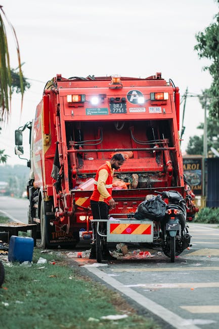 A street scene showing a waste collection vehicle, painted in red, positioned on the side of the road with its rear hatch open for waste disposal. A worker dressed in a yellow and red high-visibility vest is actively loading waste material into the vehicle, with a black plastic bag and some loose debris visible nearby. The ground around the worker and vehicle has scattered rubbish, including paper and small containers, indicating an ongoing rubbish collection process. The background features trees, utility poles, and a section of a building with signage, suggesting an urban or suburban environment, possibly in Ealing. The scene is captured in daylight with overcast skies, providing clear visibility of the equipment and activity, aligning with services such as private waste collection or rubbish removal offered by companies like Rubbish Removal Ealing, supporting the context of alternative waste handling solutions outside of municipal rubbish collection.