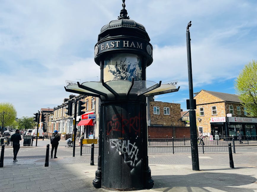 A cylindrical street clock tower with a domed black top displaying the sign 'EAST HAM' is centered in a busy urban intersection. The tower's lower section is covered in black metal mesh with graffiti tags in white and red spray paint. Surrounding the structure are traffic lights, a tall, unlit streetlamp, and several people walking along the pavement. To the right, a row of brick residential buildings with pitched roofs and a commercial shop with a pink sign are visible. The sky is partly cloudy with patches of blue, and a few green trees are seen in the background. The scene captures a typical day in East London with a focus on the street's public fixtures and urban environment, indirectly illustrating aspects of local municipal rubbish and waste management through the urban setting.
