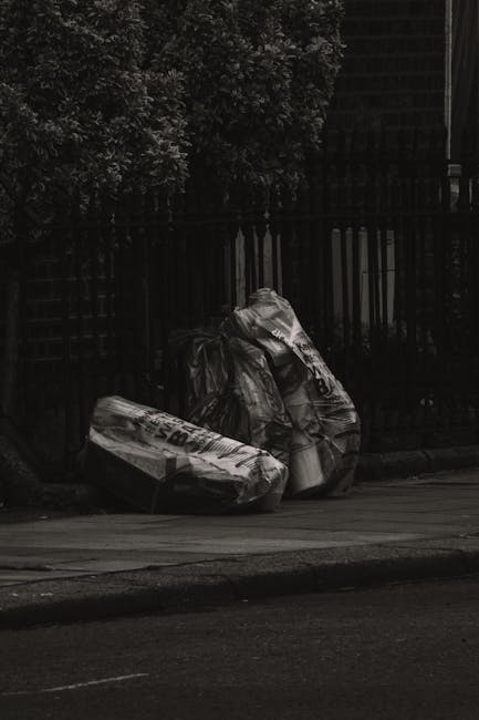 Three large black garbage bags, made of thick plastic with a glossy finish, are piled together on the edge of a paved sidewalk. The bags appear to be filled with waste, with some sections slightly bulging and tied at the top to contain their contents. They are positioned next to a black metal fence with vertical slats, which runs parallel to the curb, and behind the fence, there are dense leafy trees creating a shaded background. The scene is set outdoors in a residential or urban area, with minimal natural light illuminating the bags, casting faint shadows on the pavement. The overall environment suggests a local waste collection area, with the bags temporarily placed for rubbish removal or private disposal. Rubbish Removal Ealing might handle such waste as part of an independent rubbish clearance service, reflecting typical urban rubbish management practices.